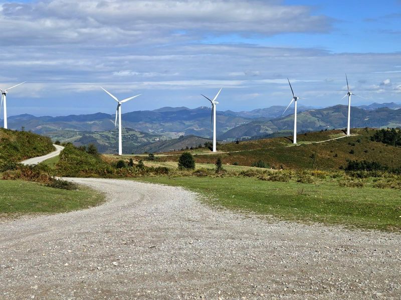 Camino de grava que se bifurca en una loma, con aerogeneradores y monta&ntilde;as al fondo