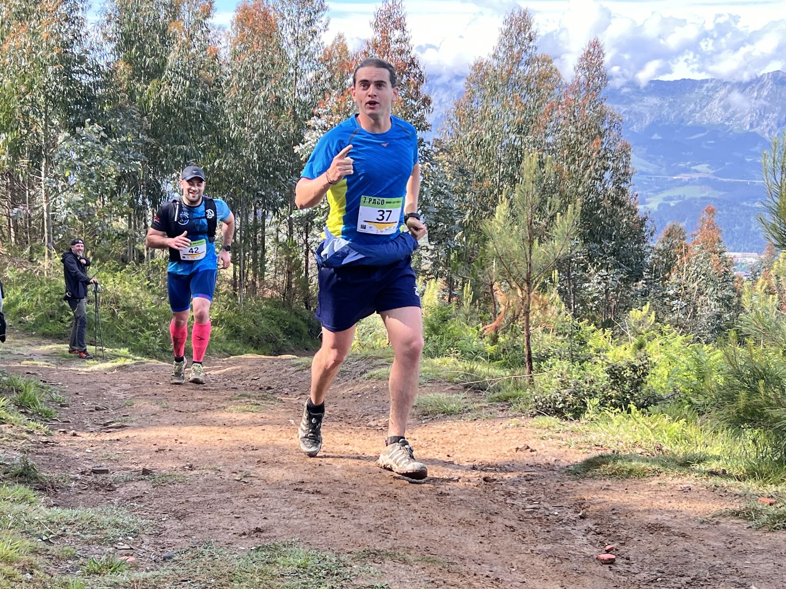 Corredor con dorsal de la 7 Pago dando el pulgar hacia arriba en un tramo de bosque, con otro corredor detr&aacute;s y las monta&ntilde;as al fondo, dorsal 37