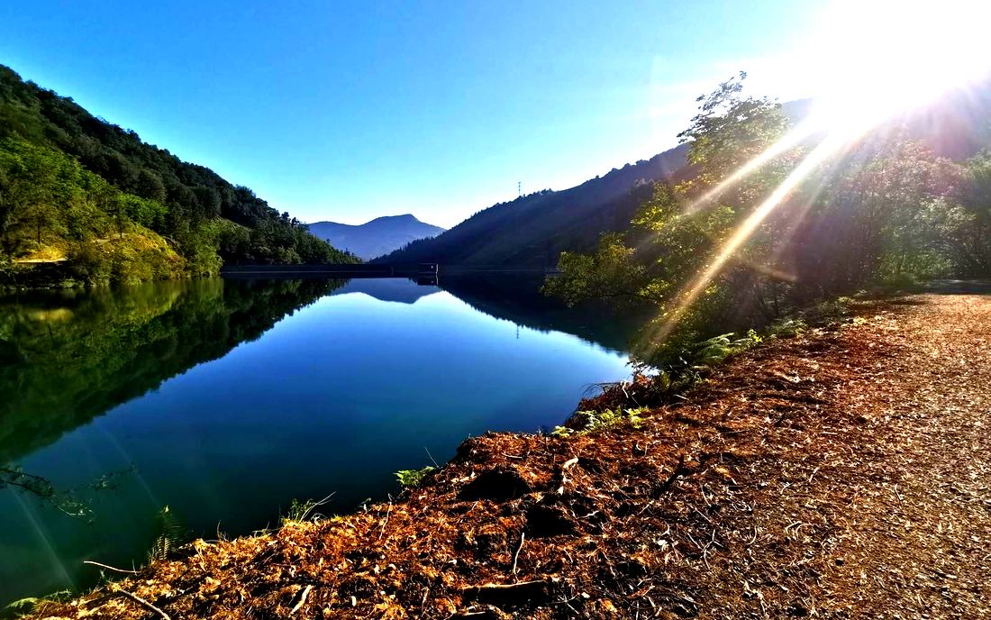 El embalse de Aixola al amanecer, con el agua en calma reflejando las laderas, el muro de la presa al fondo y el sol saliendo entre los árboles de la orilla