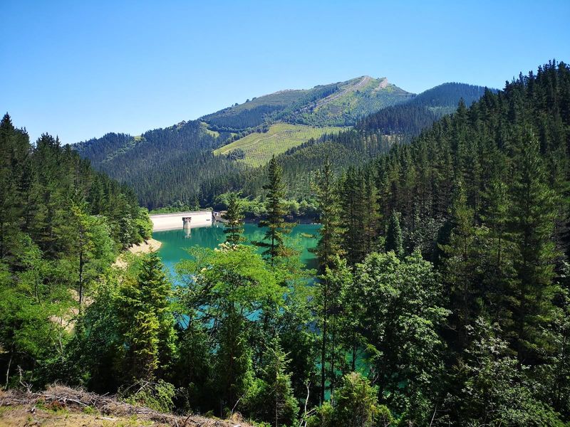El embalse de Aixola, de agua turquesa, con la presa al fondo y los montes del entorno