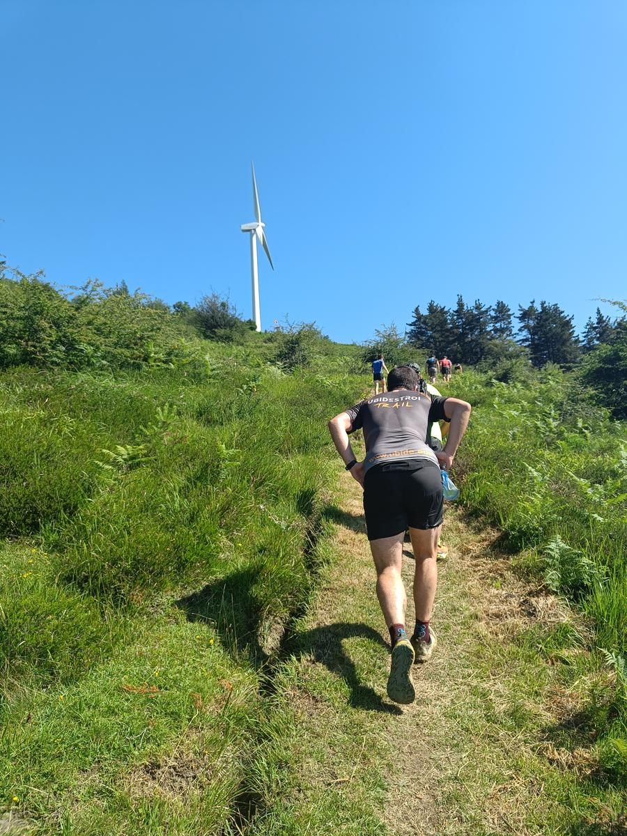 Corredor subiendo un sendero hacia un aerogenerador, con m&aacute;s corredores al fondo
