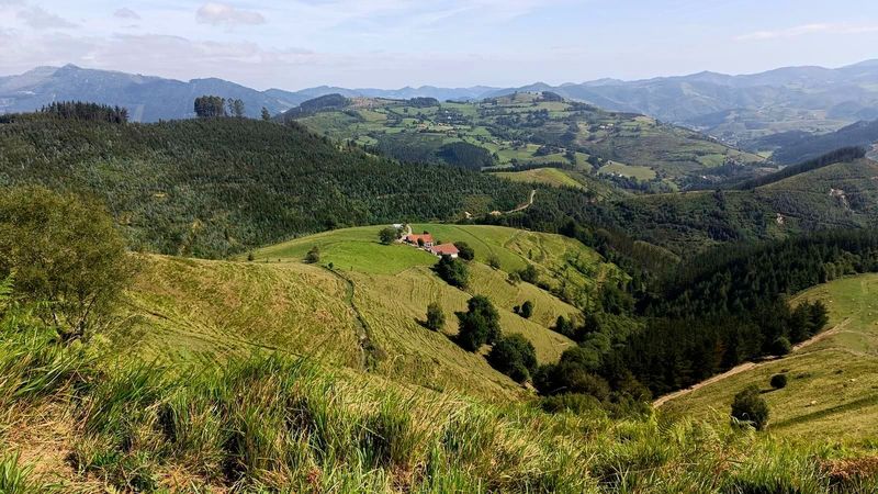 Un caser&iacute;o en lo alto de una loma, rodeado de prados y bosque, con montes al fondo