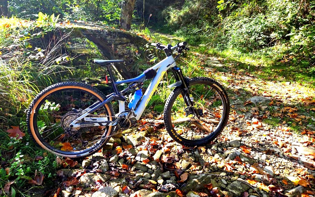 La e-bike apoyada junto a un puente de piedra de un solo arco, sobre el cauce cubierto de hojas secas