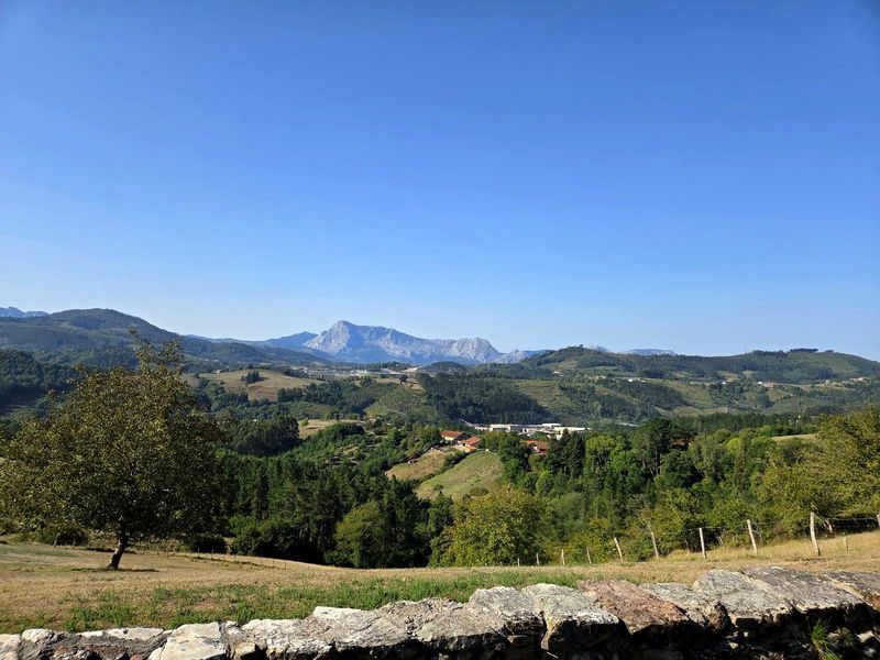 Vista del valle desde un muro de piedra, con caser&iacute;os entre prados y bosque y una sierra rocosa al fondo