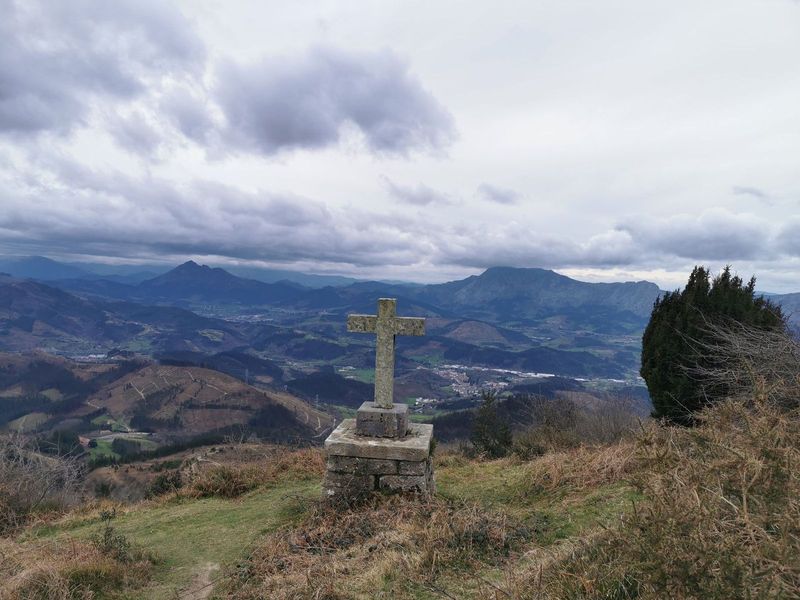 Cruz de piedra en la cima, con el valle y varios pueblos al fondo entre nubes bajas