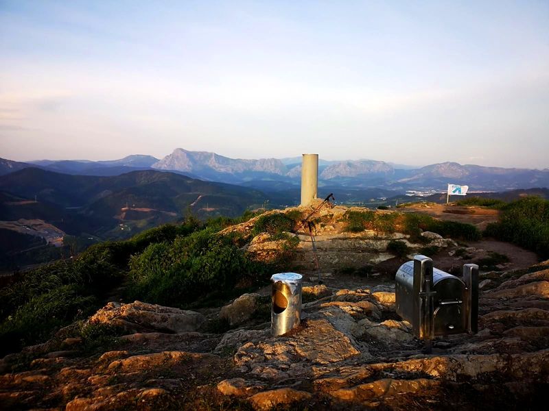 V&eacute;rtice geod&eacute;sico en la cumbre del Urko, con bastones de trekking apoyados y las monta&ntilde;as del entorno al fondo