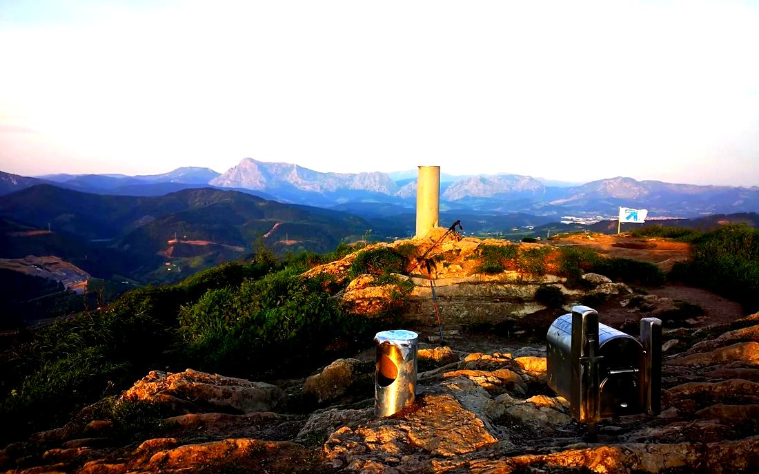 Vértice geodésico en la cumbre del Urko, con bastones de trekking apoyados y las montañas del entorno al fondo