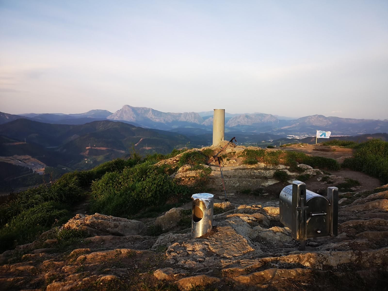 Vértice geodésico en la cumbre del Urko, con bastones de trekking apoyados y las montañas del entorno al fondo