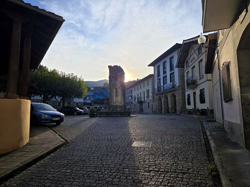 Bolibarko plaza egunsentian, harrizko monumentu bat erdian eta eguzkia eraikinen artetik agertzen