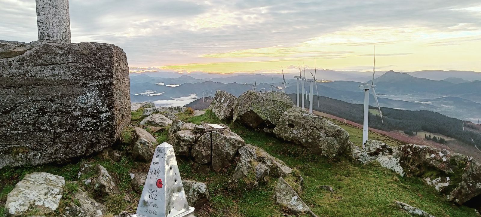 Oizko gailurreko mugarria (1.026 m) harrizko gurutze baten ondoan, aerosorgailuak eta mendiak ilunabarrean