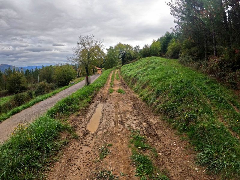 Pista de tierra embarrada subiendo junto al asfalto, con un moj&oacute;n y los montes al fondo