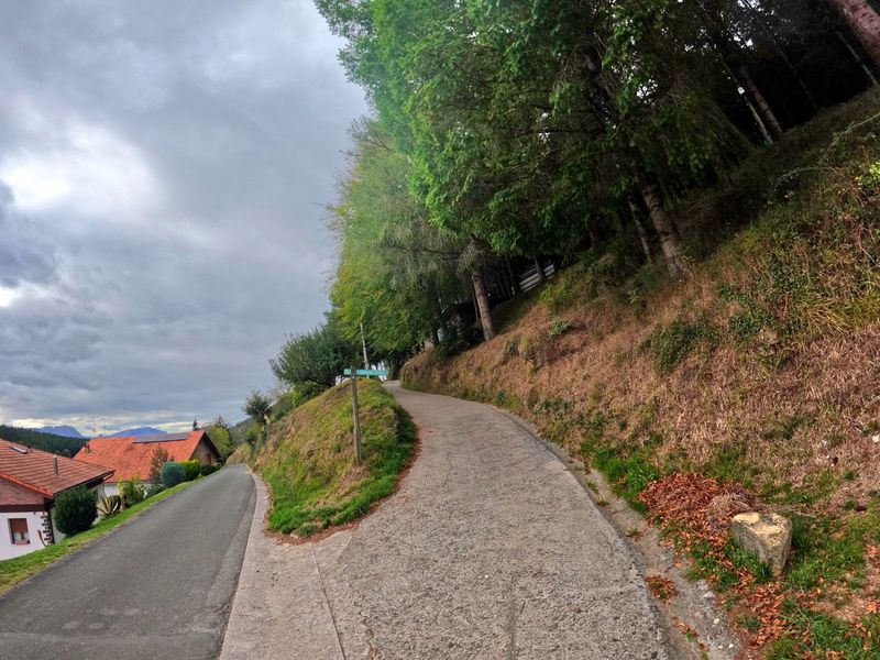 Pista de cemento subiendo junto a los caser&iacute;os, con el cartel del barrio y los montes al fondo