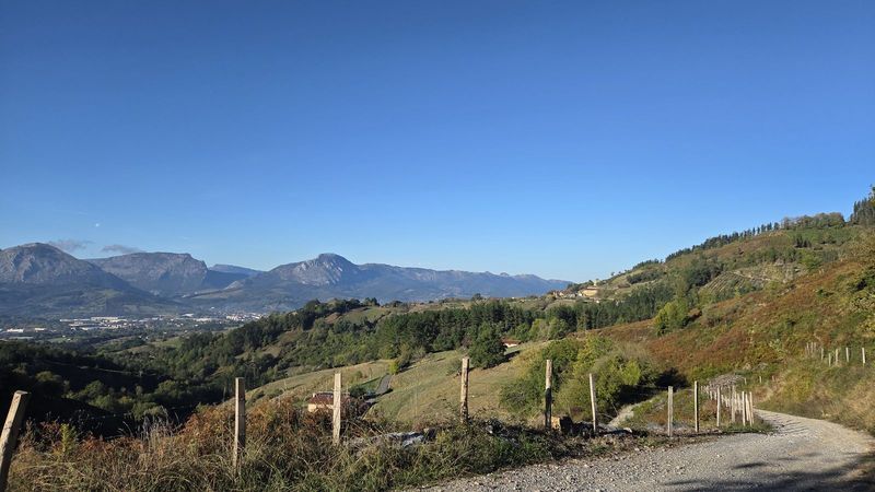 Pista de piedra en alto sobre el valle, con prados y caser&iacute;os en la ladera y los montes al fondo