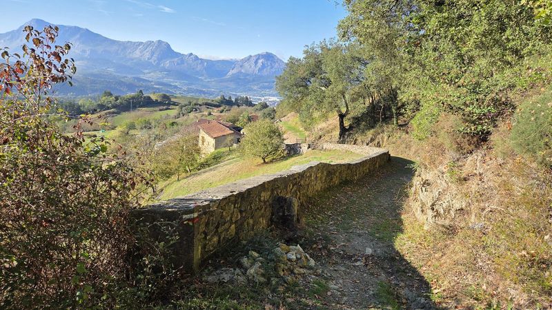 Camino con muro de piedra sobre un caser&iacute;o, con las monta&ntilde;as al fondo