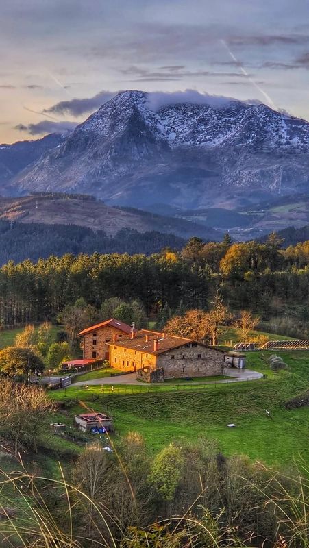 Caser&iacute;o tradicional junto a un bosque, con un monte nevado al fondo, al atardecer