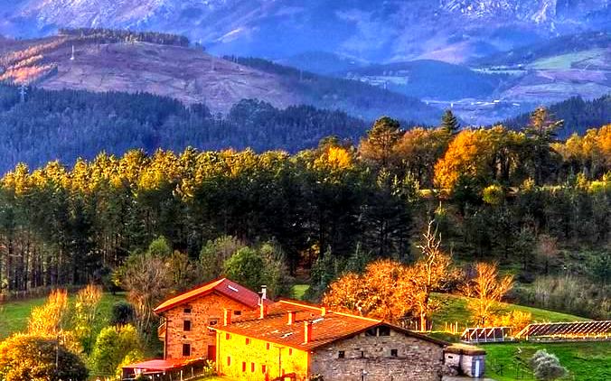 Caserío tradicional junto a un bosque, con un monte nevado al fondo, al atardecer