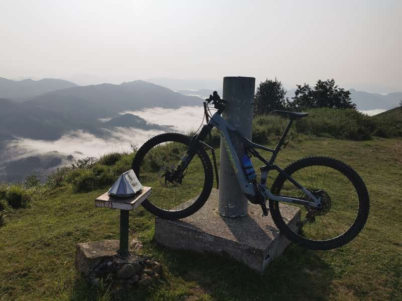 Bicicleta el&eacute;ctrica apoyada en el v&eacute;rtice geod&eacute;sico de la cima, sobre un mar de nubes con monta&ntilde;as asomando al fondo