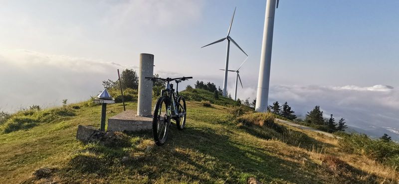 Bicicleta apoyada en un moj&oacute;n de piedra junto a aerogeneradores, sobre un mar de nubes
