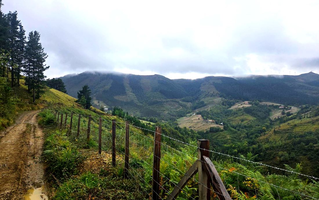 Vista de un valle con una alambrada de madera en primer plano, montes con aerogeneradores entre las nubes y un caserío disperso al fondo