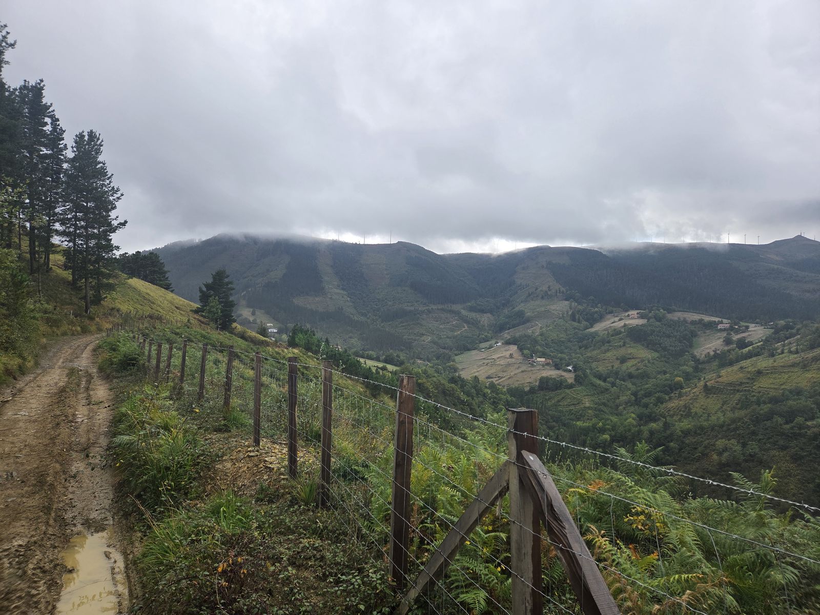 Vista de un valle con una alambrada de madera en primer plano, montes con aerogeneradores entre las nubes y un caser&iacute;o disperso al fondo