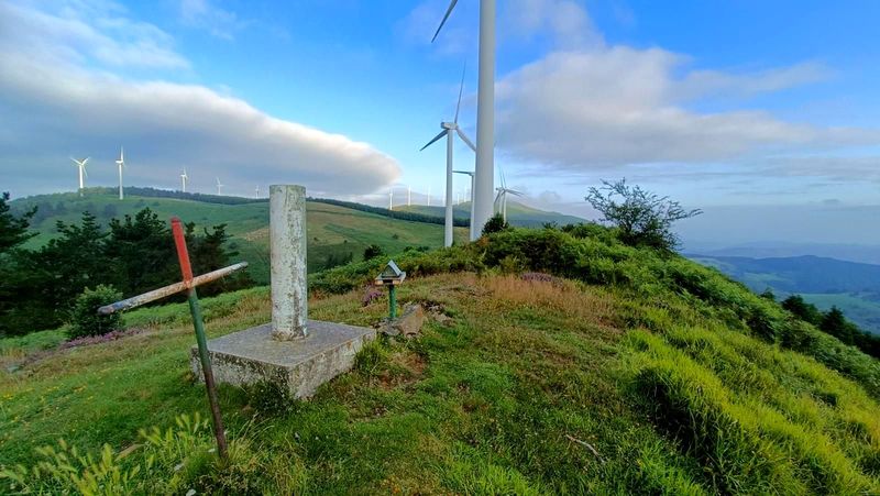 Cruz de hierro y v&eacute;rtice geod&eacute;sico en una cima, con aerogeneradores cerca y un banco de nubes al fondo