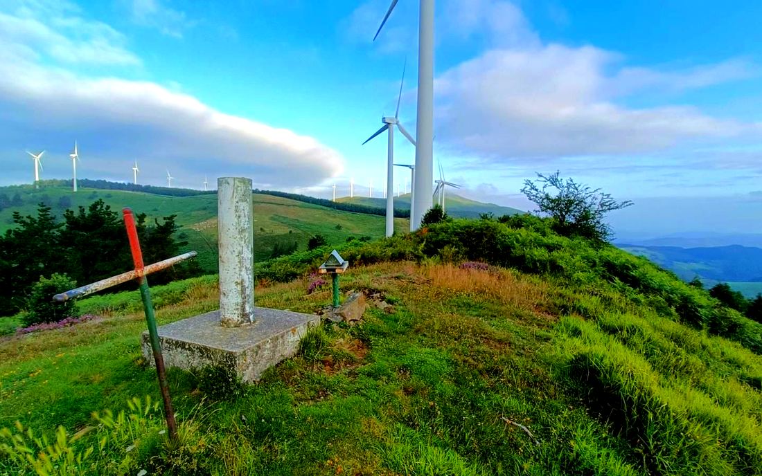 Cruz de hierro y vértice geodésico en una cima, con aerogeneradores cerca y un banco de nubes al fondo