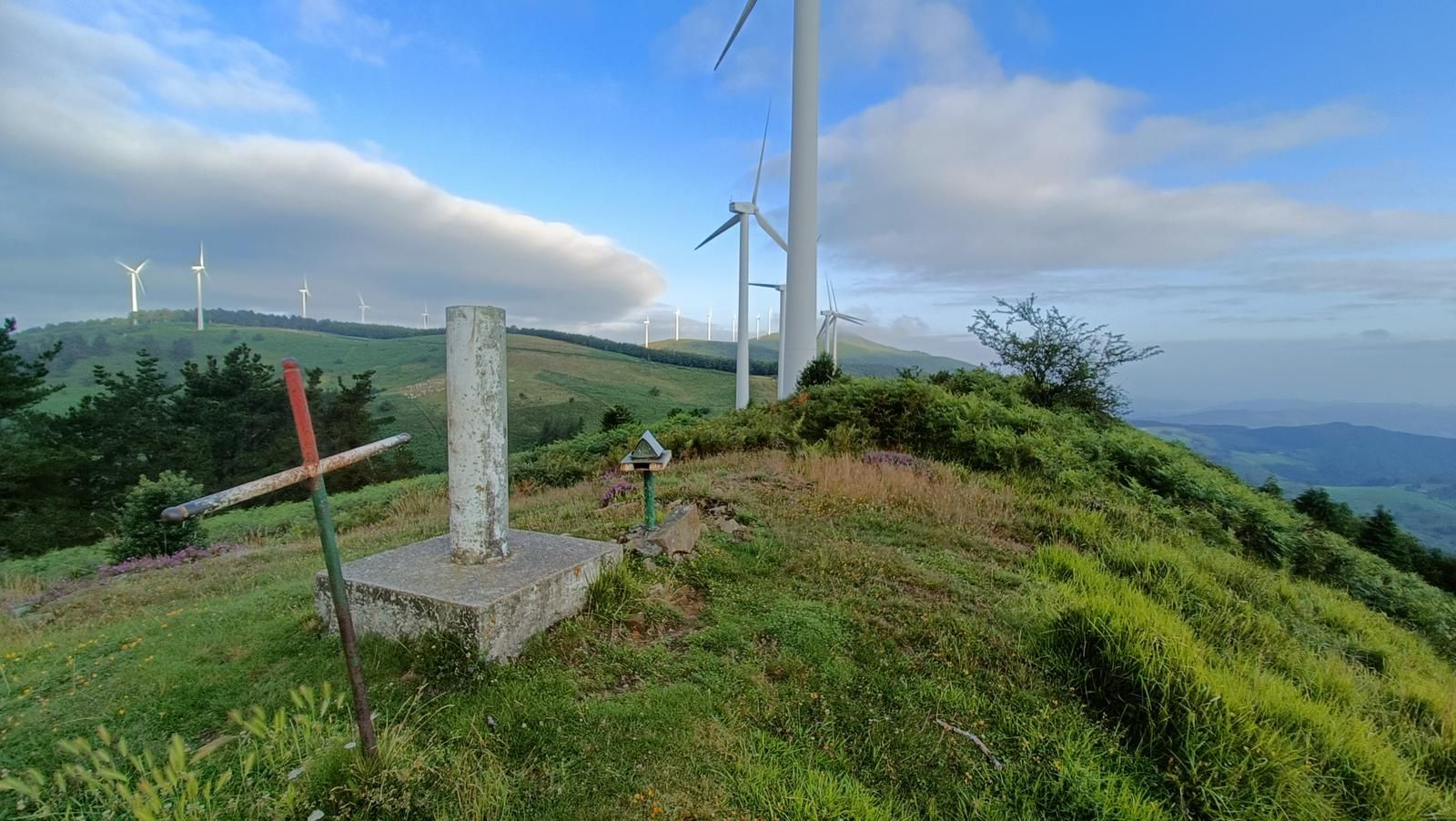 Cruz de hierro y v&eacute;rtice geod&eacute;sico en una cima, con aerogeneradores cerca y un banco de nubes al fondo