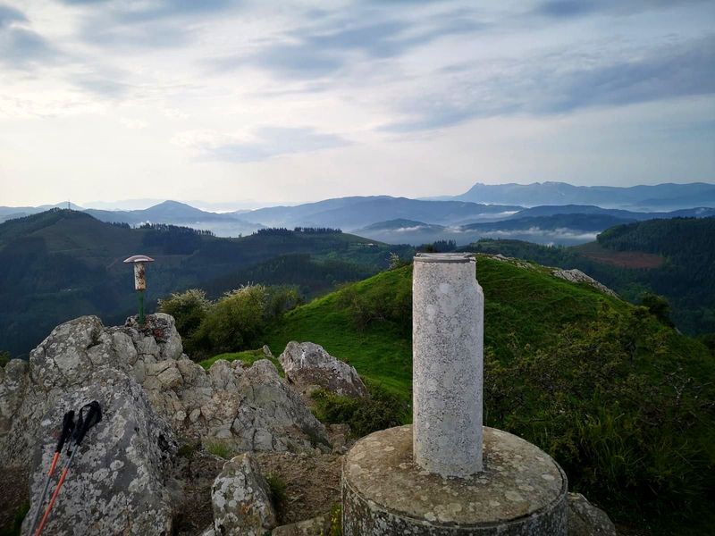 V&eacute;rtice geod&eacute;sico en una cima, con bastones de trekking apoyados en las rocas y monta&ntilde;as al fondo