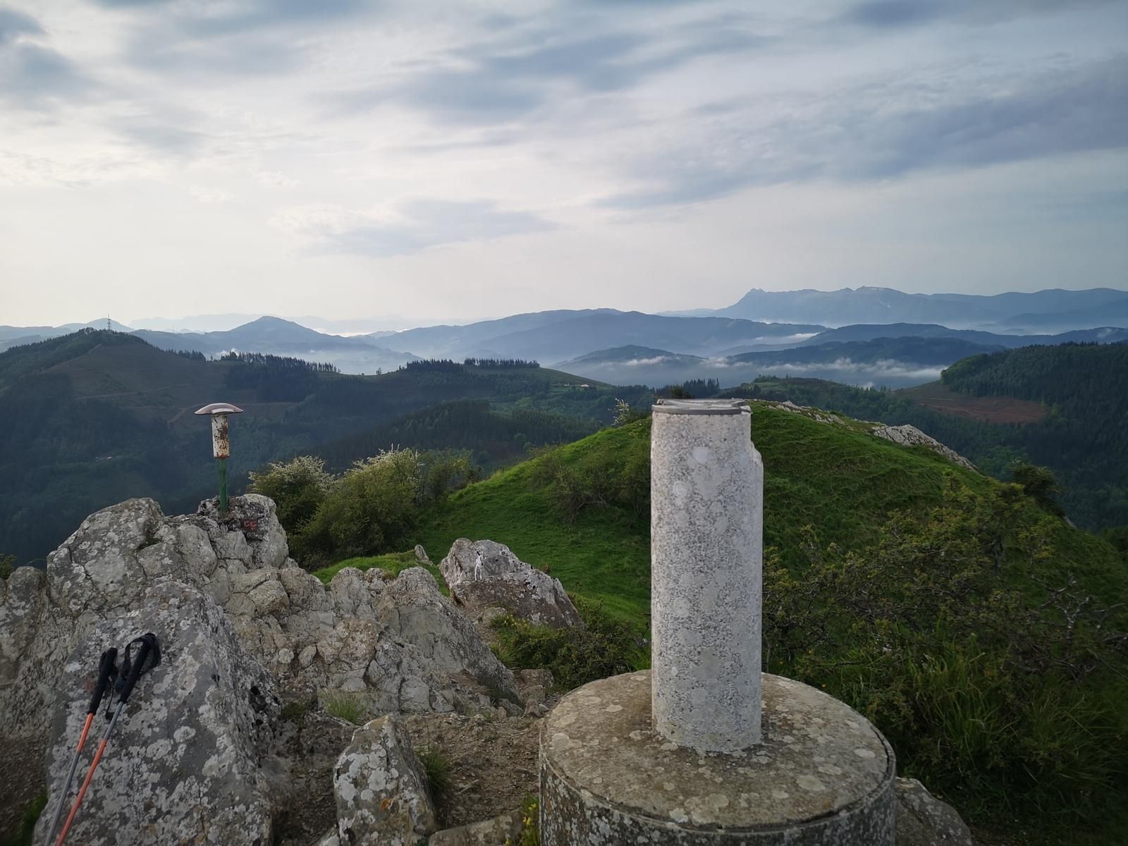 V&eacute;rtice geod&eacute;sico en una cima, con bastones de trekking apoyados en las rocas y monta&ntilde;as al fondo