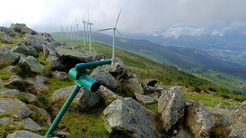 Baliza verde de sendero entre rocas, con una hilera de aerogeneradores en la cresta y el valle al fondo