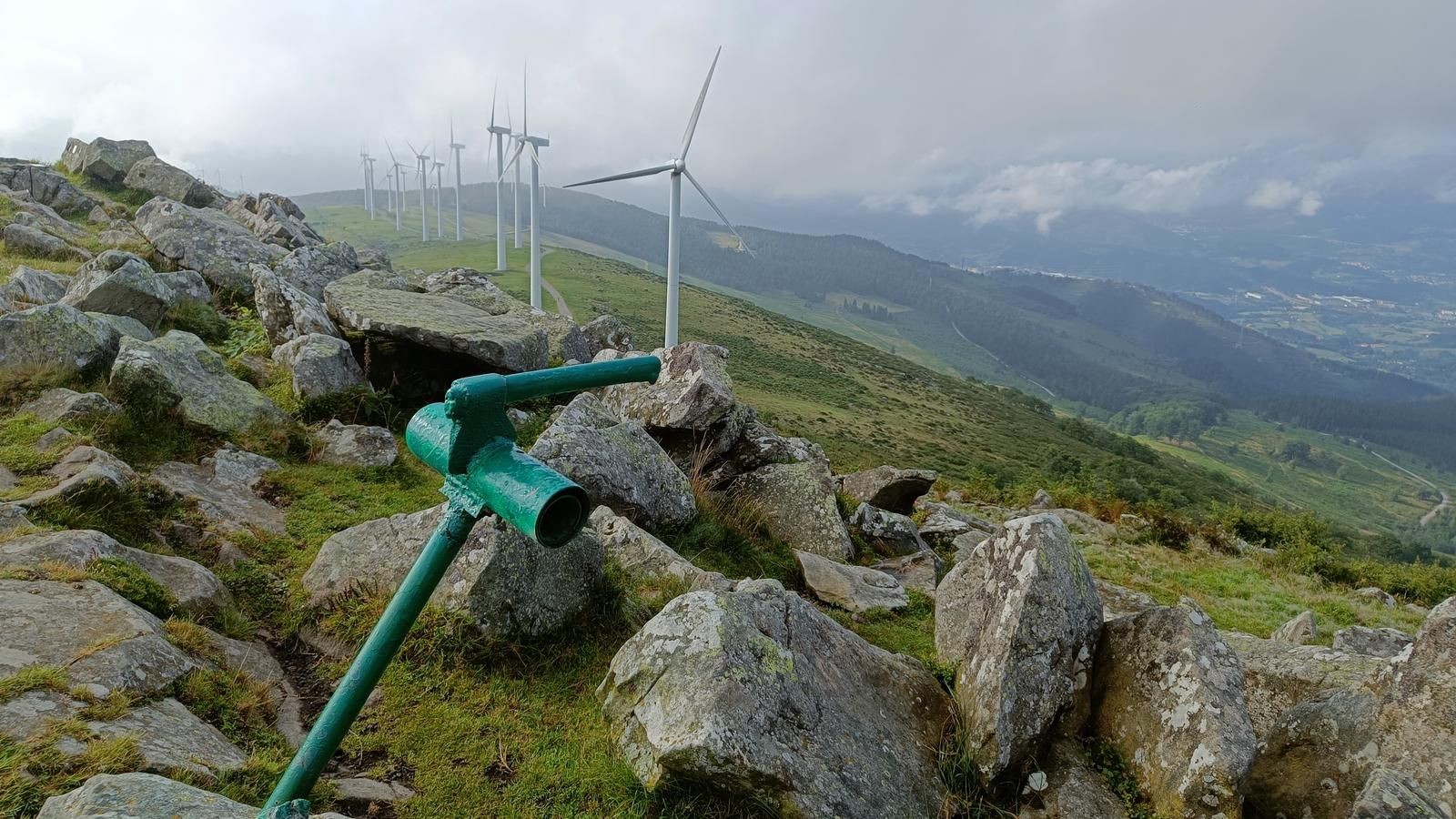 Baliza verde de sendero entre rocas, con una hilera de aerogeneradores en la cresta y el valle al fondo