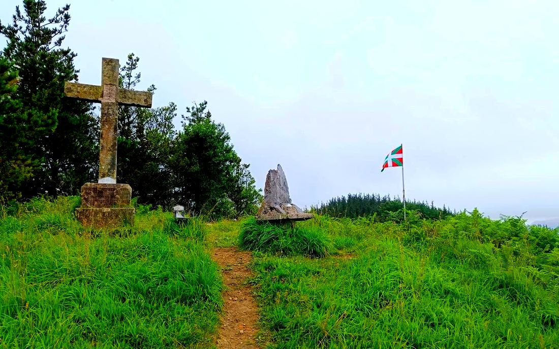 Cruz de piedra sobre su pedestal en lo alto, junto a un monolito de roca y una ikurriña ondeando al fondo, entre hierba alta y pinar