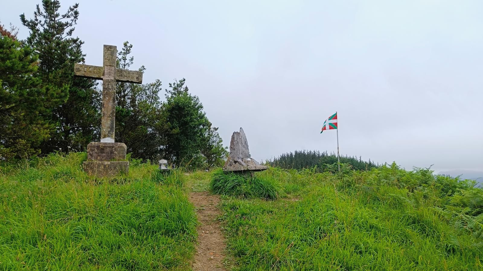 Cruz de piedra sobre su pedestal en lo alto, junto a un monolito de roca y una ikurri&ntilde;a ondeando al fondo, entre hierba alta y pinar
