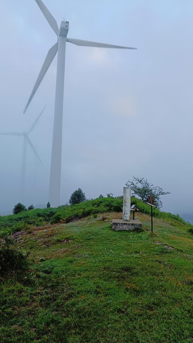 Aerogenerador entre la niebla sobre una loma de hierba, con un monolito de piedra y una cruz met&aacute;lica en primer plano