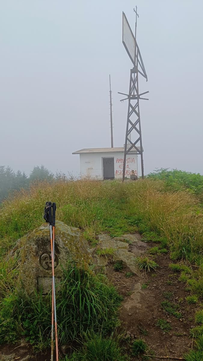 La cima de Intxorta entre la niebla, con su caseta pintada y la torreta met&aacute;lica, una piedra tallada con un lauburu y unos bastones apoyados en primer plano