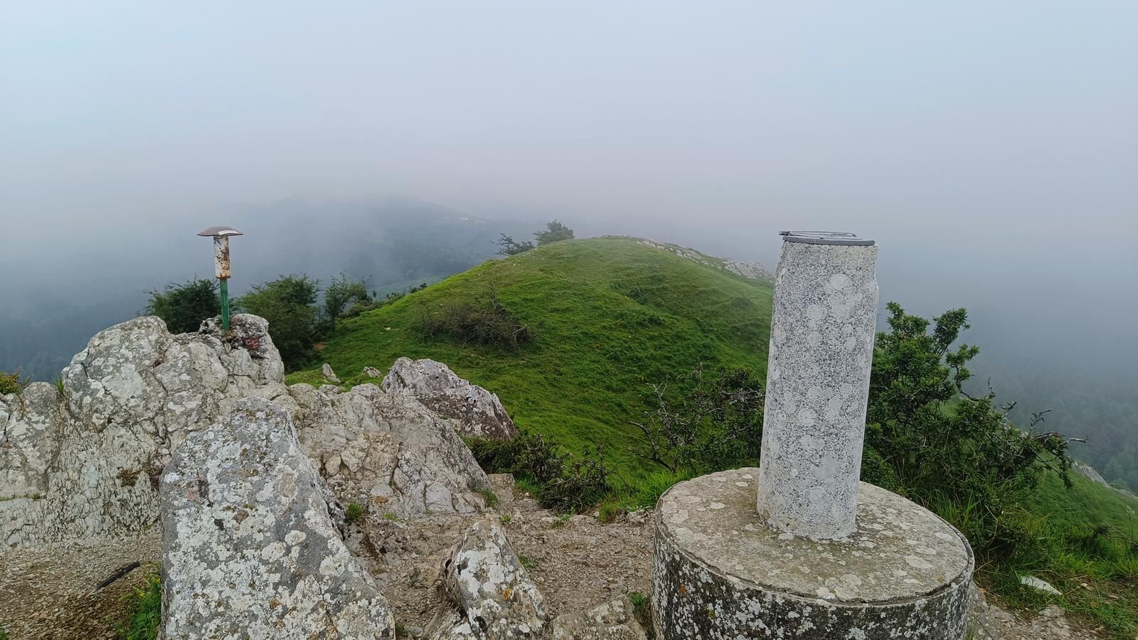 V&eacute;rtice geod&eacute;sico sobre su base de hormig&oacute;n en la cima, entre roca caliza, con el cordal de hierba perdi&eacute;ndose en la niebla