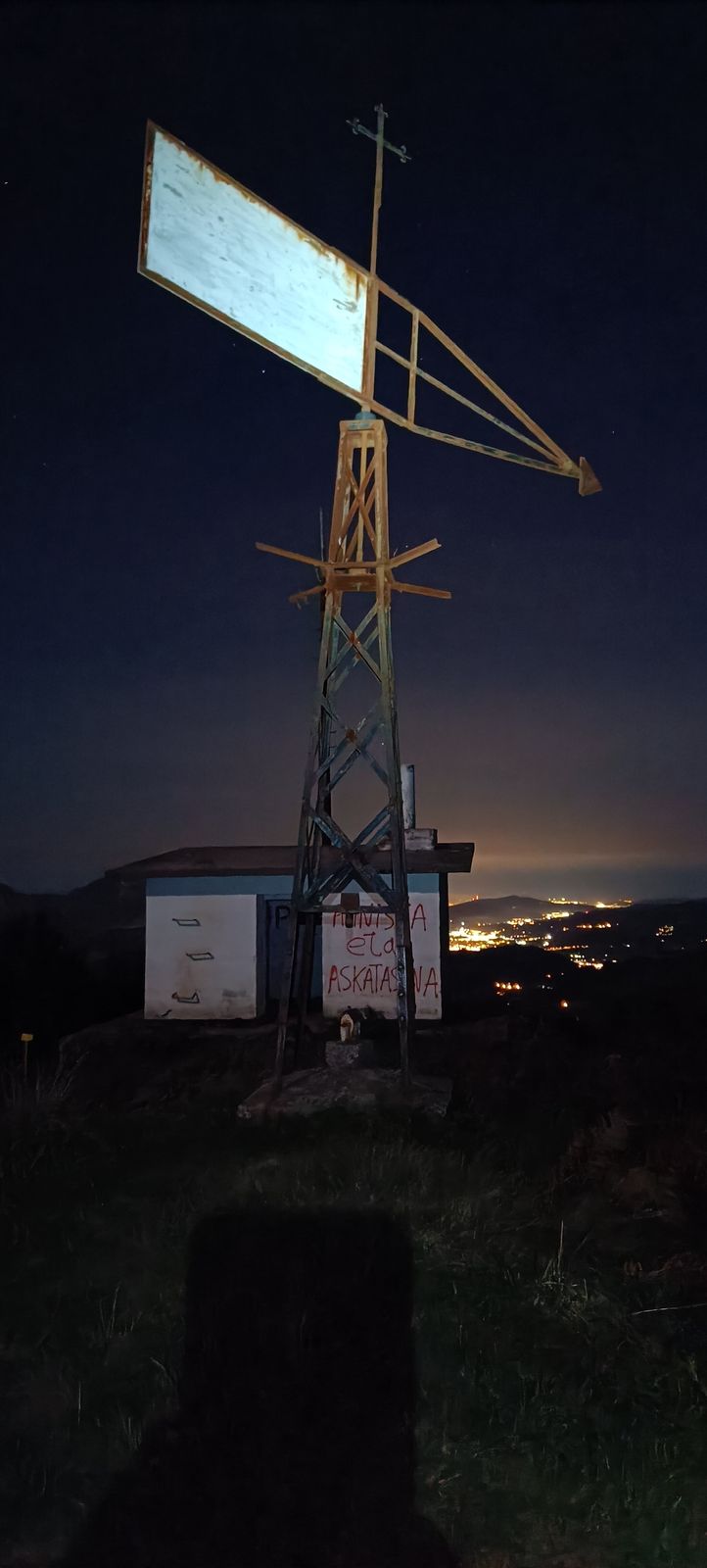 Intxorta de noche: la torreta met&aacute;lica con su paleta y la caseta con pintadas, con las luces de los pueblos del valle al fondo