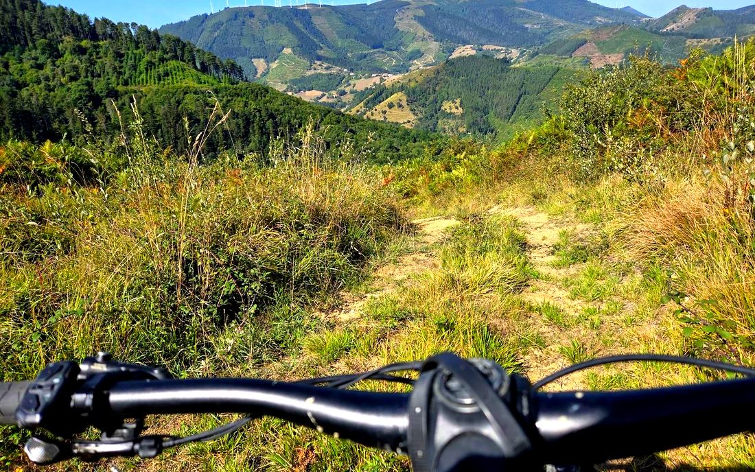 Manillar de la bicicleta eléctrica en un sendero entre matorral, con los aerogeneradores del Oiz al fondo