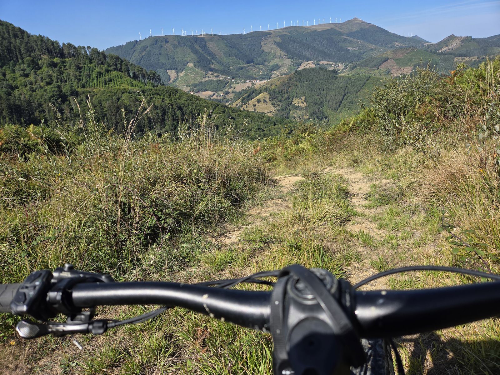 Manillar de la bicicleta eléctrica en un sendero entre matorral, con los aerogeneradores del Oiz al fondo