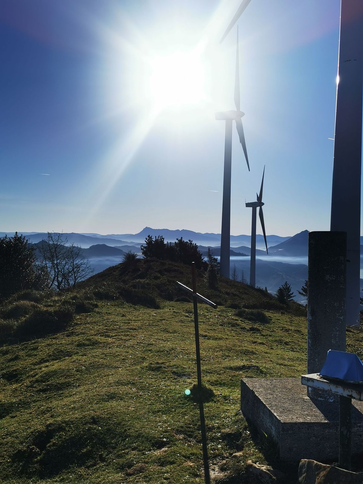 Aerogeneradores del parque e&oacute;lico recortados contra el sol, con la niebla en los valles al fondo