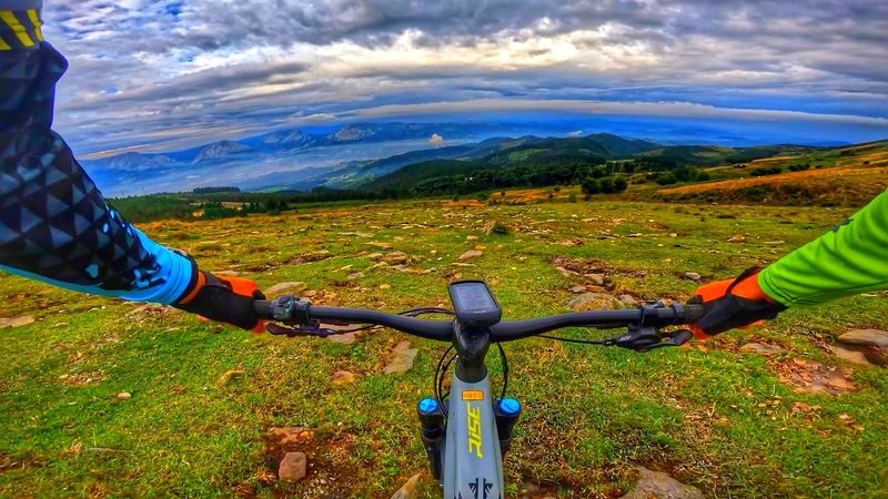 E-bikearen eskulekutik ikusitako larrea, harriekin eta mendiak atzealdean, zeru hodeitsu baten azpian