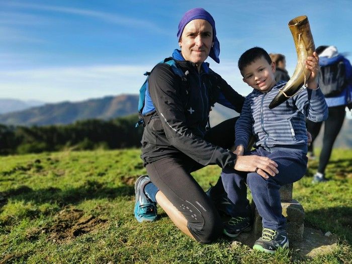Heldu bat eta haur bat ardo-zahato batekin posatzen Mendibilgo gailurrean, inguruko mendiak atzealdean