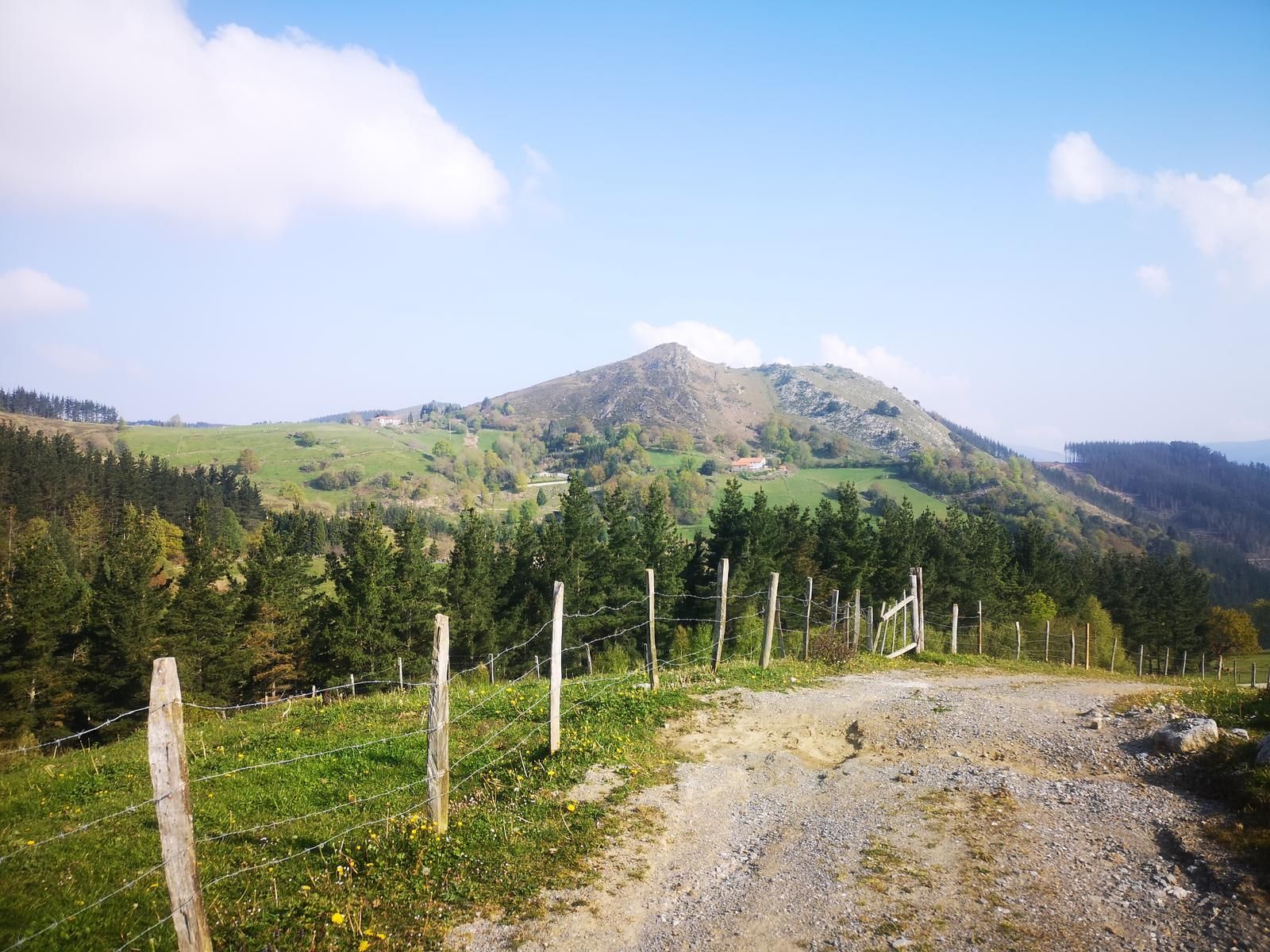 Pico rocoso al fondo, visto desde un camino de tierra junto a una alambrada, con caser&iacute;os dispersos en la ladera