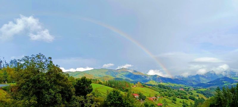 Arco&iacute;ris sobre un valle verde con caser&iacute;os de tejado rojo y montes al fondo