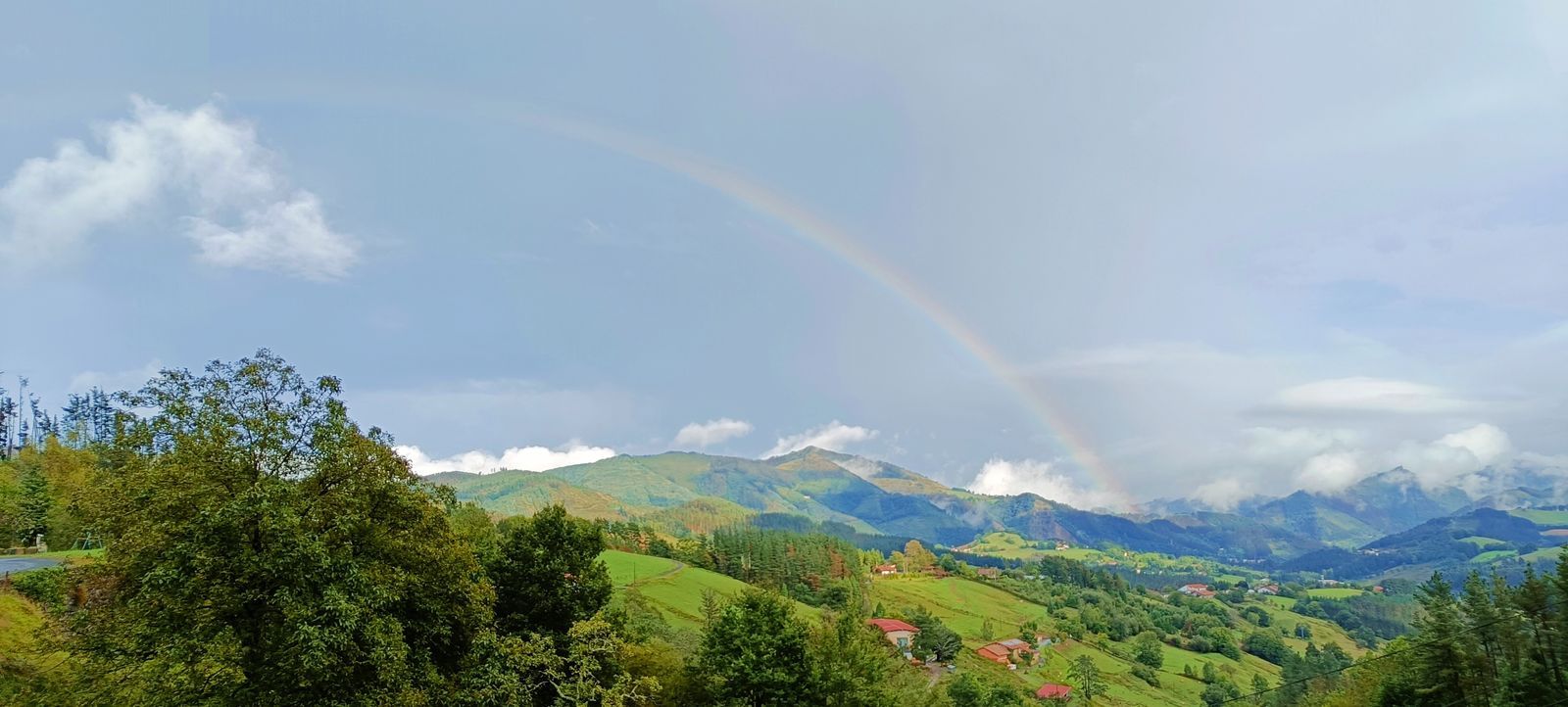 Arco&iacute;ris sobre un valle verde con caser&iacute;os de tejado rojo y montes al fondo