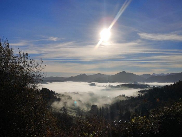 Vista de un valle cubierto de niebla desde lo alto, con el sol asomando entre los &aacute;rboles y montes boscosos alrededor