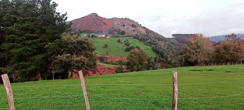 Astarloko Atxa, el picacho de roca rojiza sobre Muniozguren, con un caser&iacute;o de tejado rojo a media ladera, prados y arbolado por debajo y un cierre de alambre en primer plano
