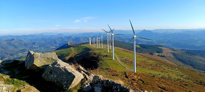 La hilera de aerogeneradores del parque e&oacute;lico recorriendo el cordal del Oiz, con grandes rocas en primer plano, ganado en los pastos y las monta&ntilde;as del entorno al fondo