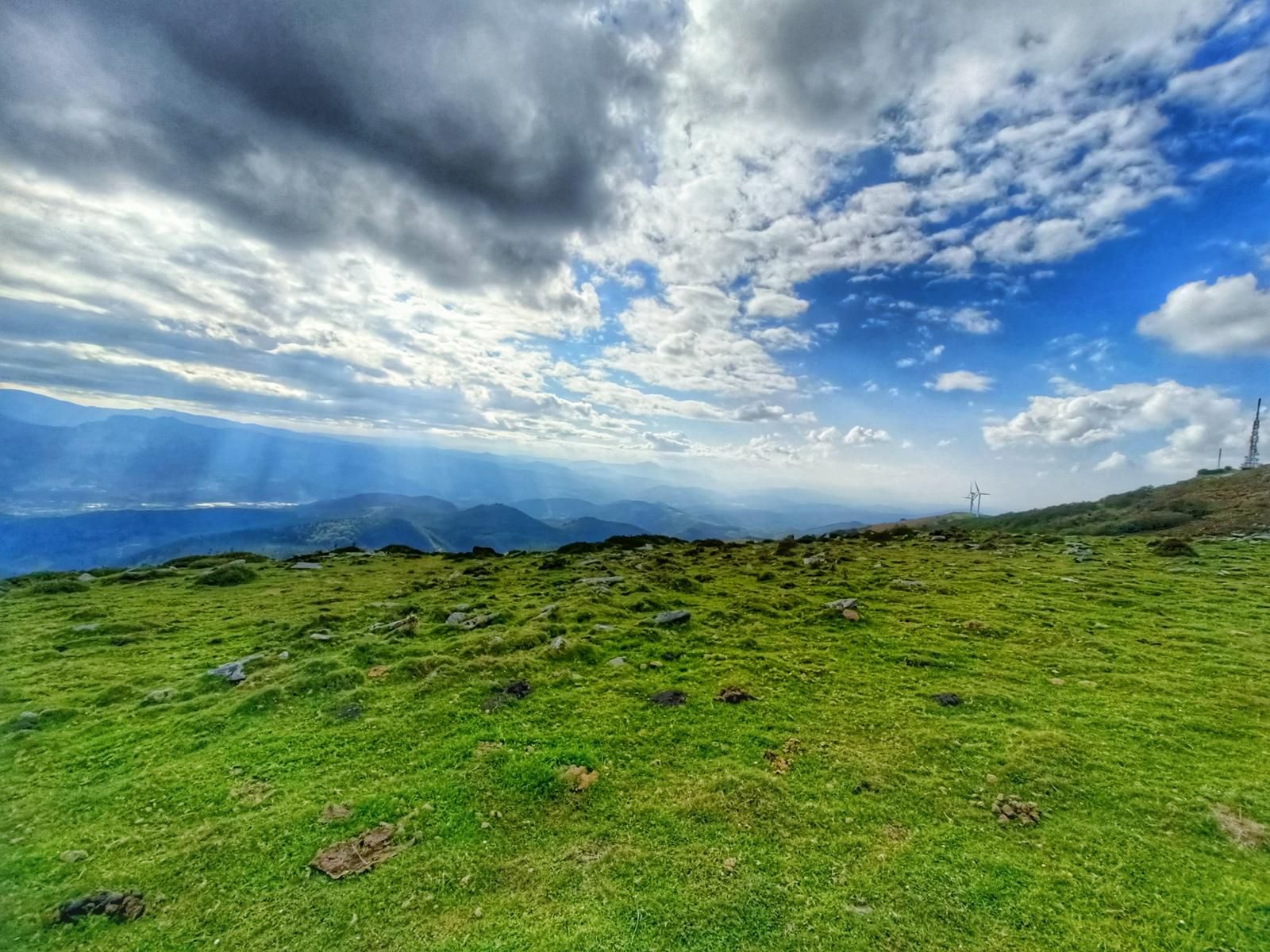 Campa de hierba en lo alto, con rayos de sol entre las nubes sobre los valles y montes del entorno