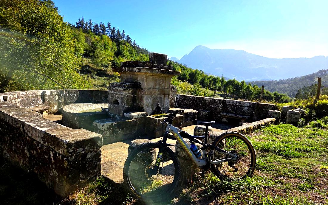 Fuente y lavadero de piedra con la bici apoyada en el muro, con una montaña al fondo
